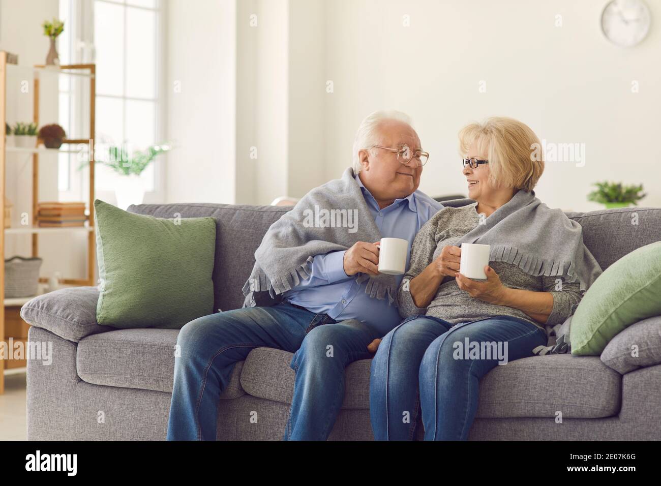 Una coppia sorridente di nonni maturi seduti insieme sul divano e riscaldandosi con tè caldo Foto Stock