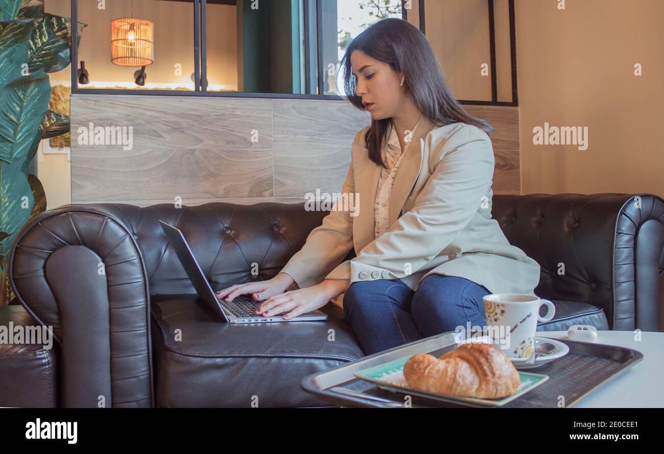 La giovane donna scrive e invia un'e-mail seduta sul divano in un bar la mattina presto mentre si serve la colazione. Concetto di Boss ragazza 2021. Foto Stock