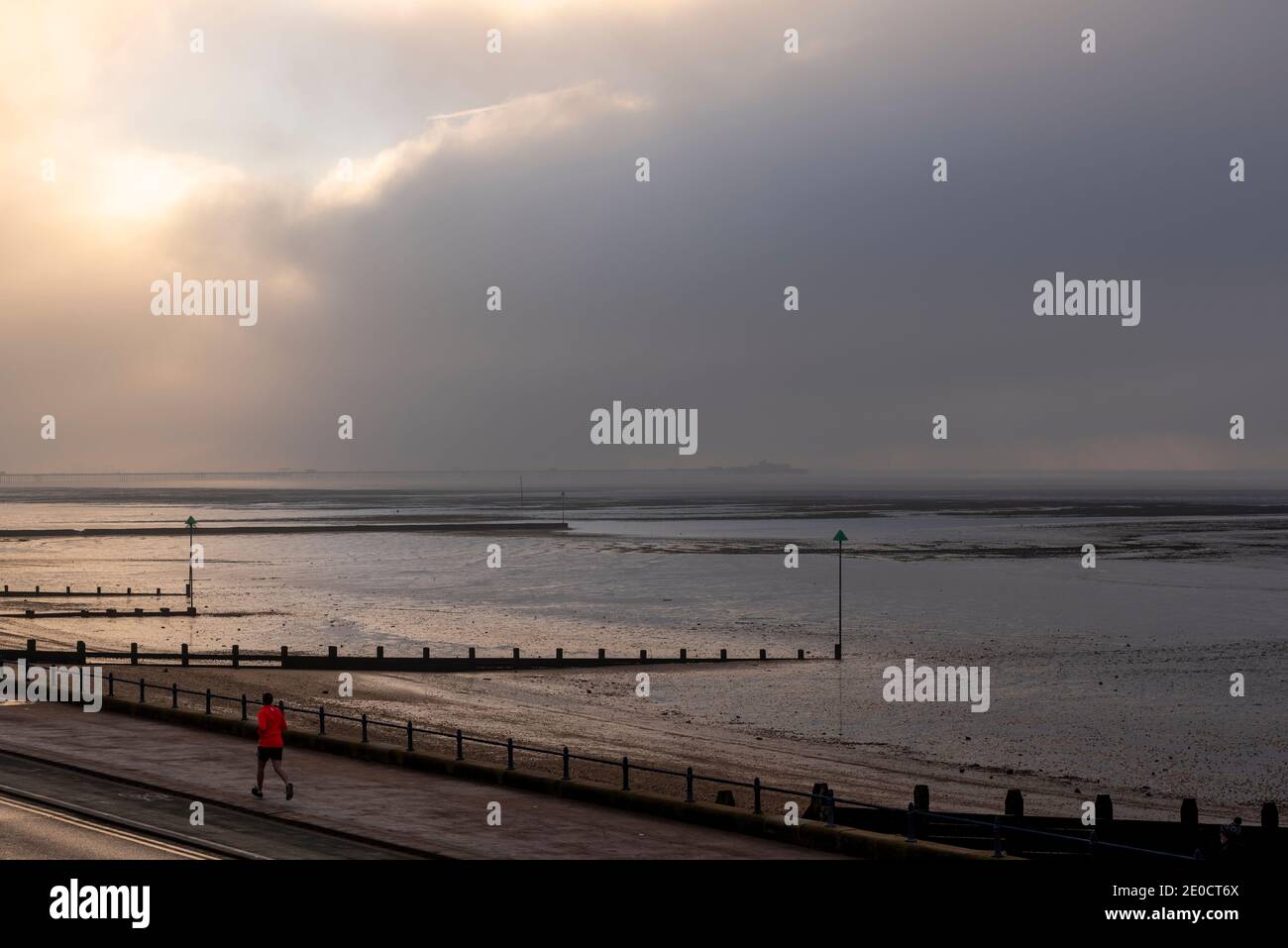 Jogger in giacca di colore brillante che corre lungo la passeggiata lungomare a Southend on Sea, Essex, Regno Unito, con il fronte meteo nero in movimento. Previsione neve Foto Stock