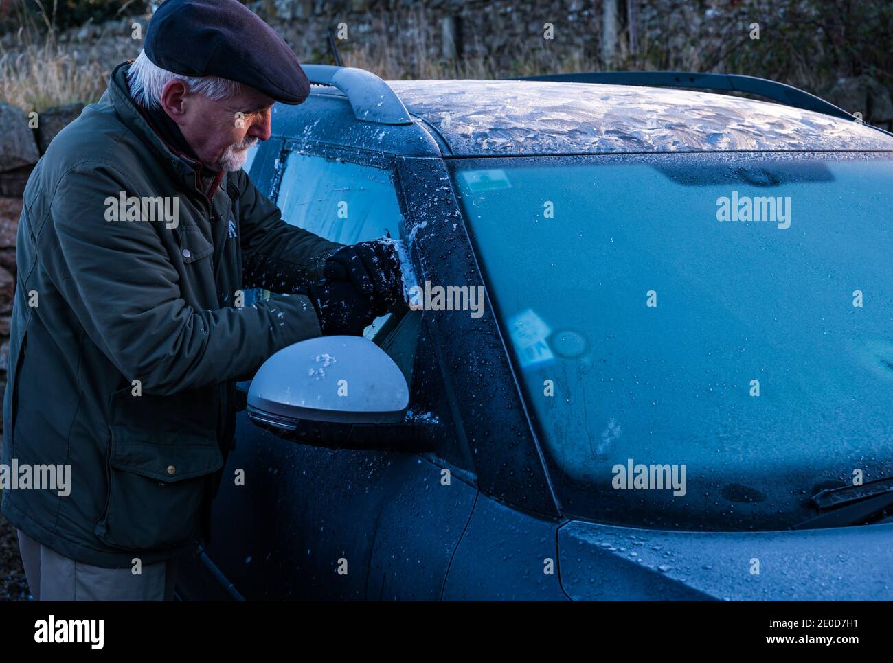 Uomo anziano raschiando il ghiaccio dal parabrezza dell'automobile in inverno con i modelli di gelo sul tetto dell'automobile, Scozia, Regno Unito Foto Stock