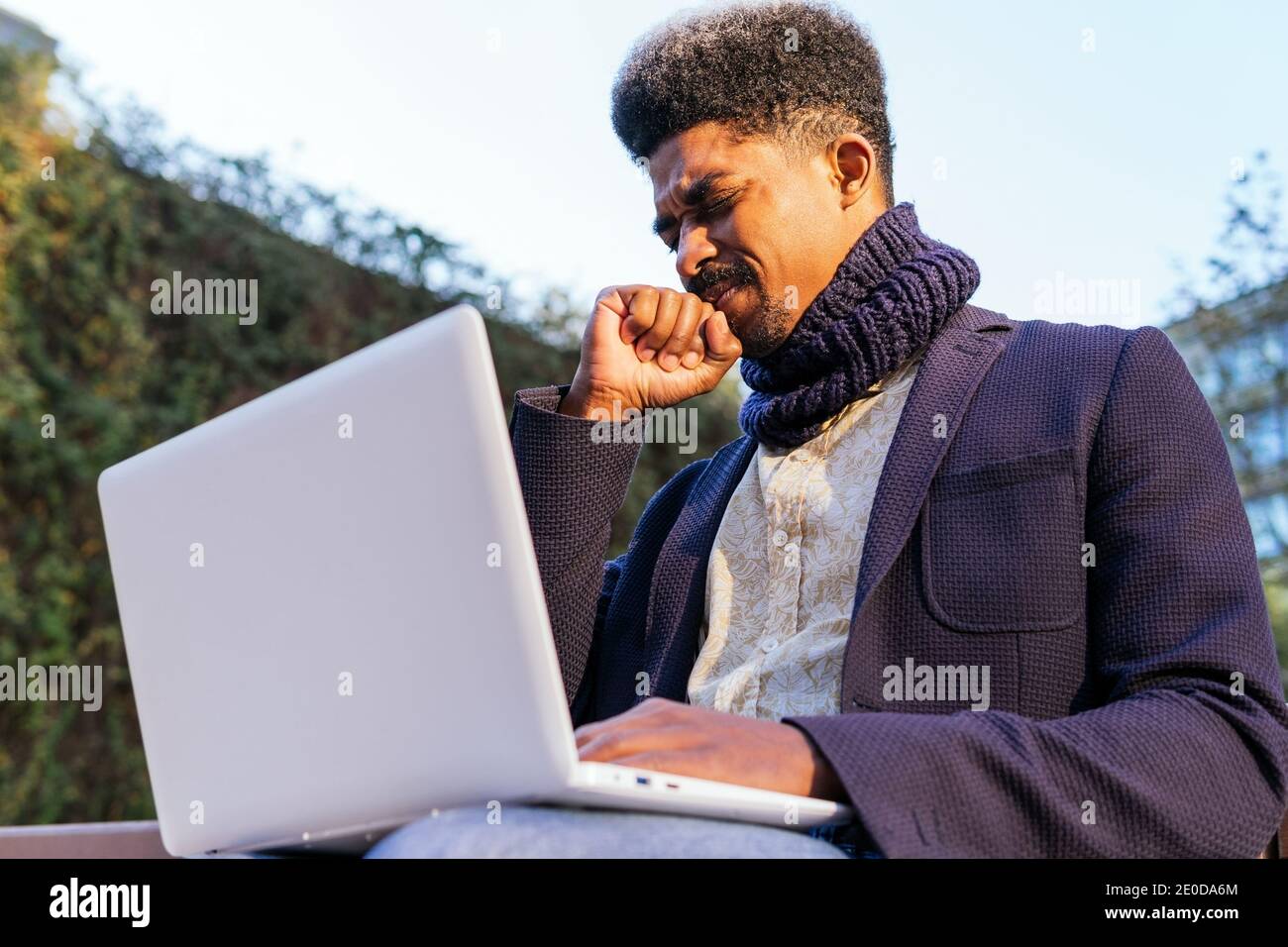 Basso angolo di imprenditore africano americano maschile seduto sulla strada con occhi chiusi e pugnalati mentre si lavora su un computer portatile e in attesa di approvazione Foto Stock