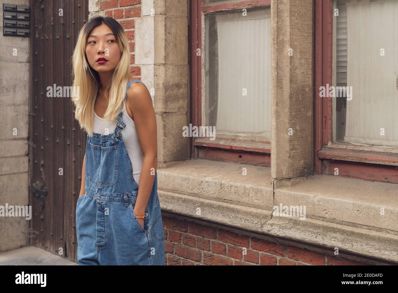 Donna asiatica sicura indossando un denim trendy in piedi con le mani in tasca in città e guardando via Foto Stock