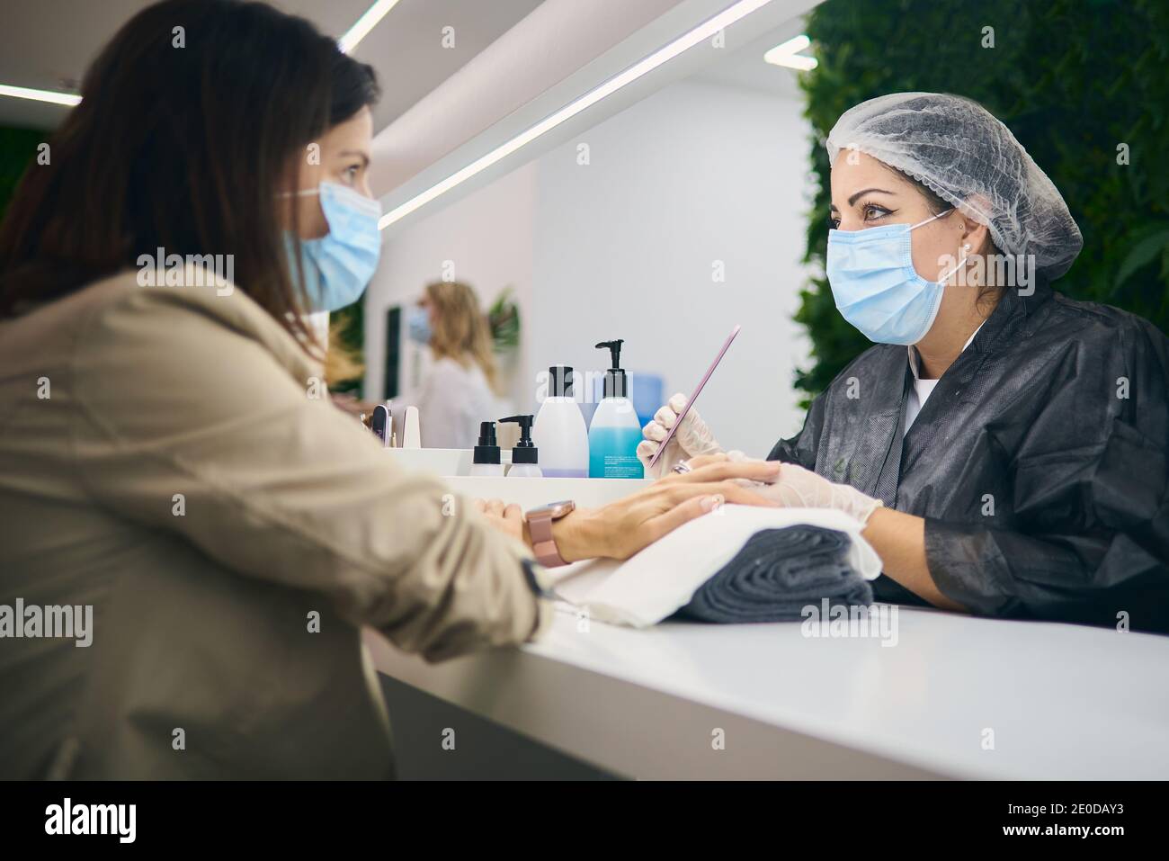 Vista laterale del maestro allegro manicure utilizzando lima unghie mentre seduto al tavolo con una cliente femminile in un moderno salone di bellezza durante la pandemia del coronavirus Foto Stock
