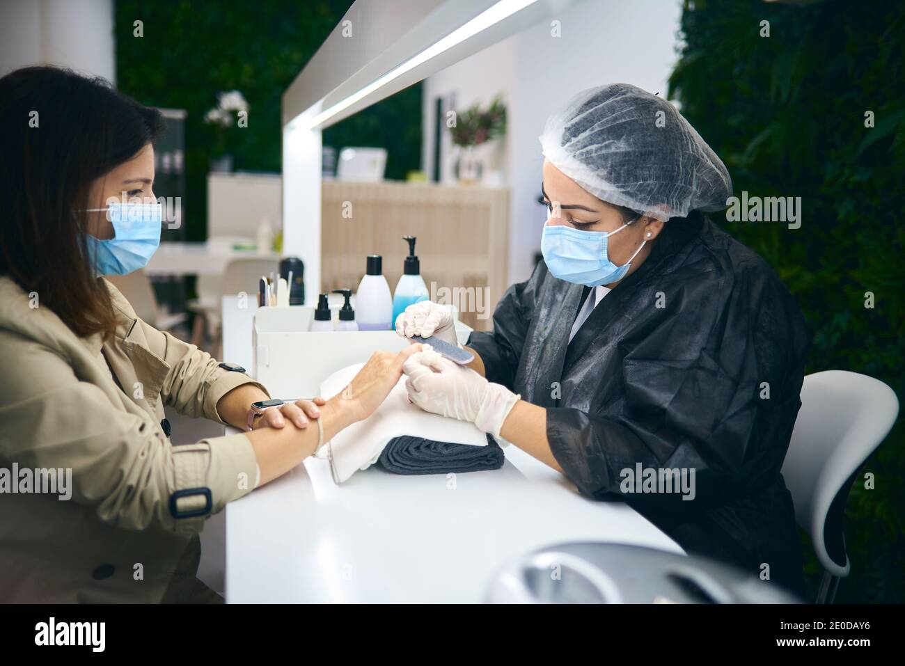 Vista laterale del maestro allegro manicure utilizzando lima unghie mentre seduto al tavolo con una cliente femminile in un moderno salone di bellezza durante la pandemia del coronavirus Foto Stock