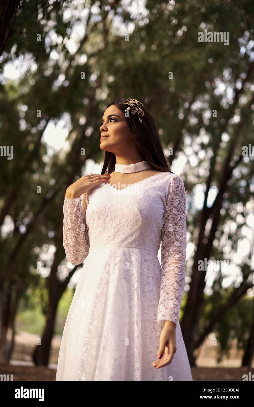 Vista laterale di elegante giovane femmina in boho bridal maxi vestitevi e cavatoie in boschi verdi e guardate via Foto Stock