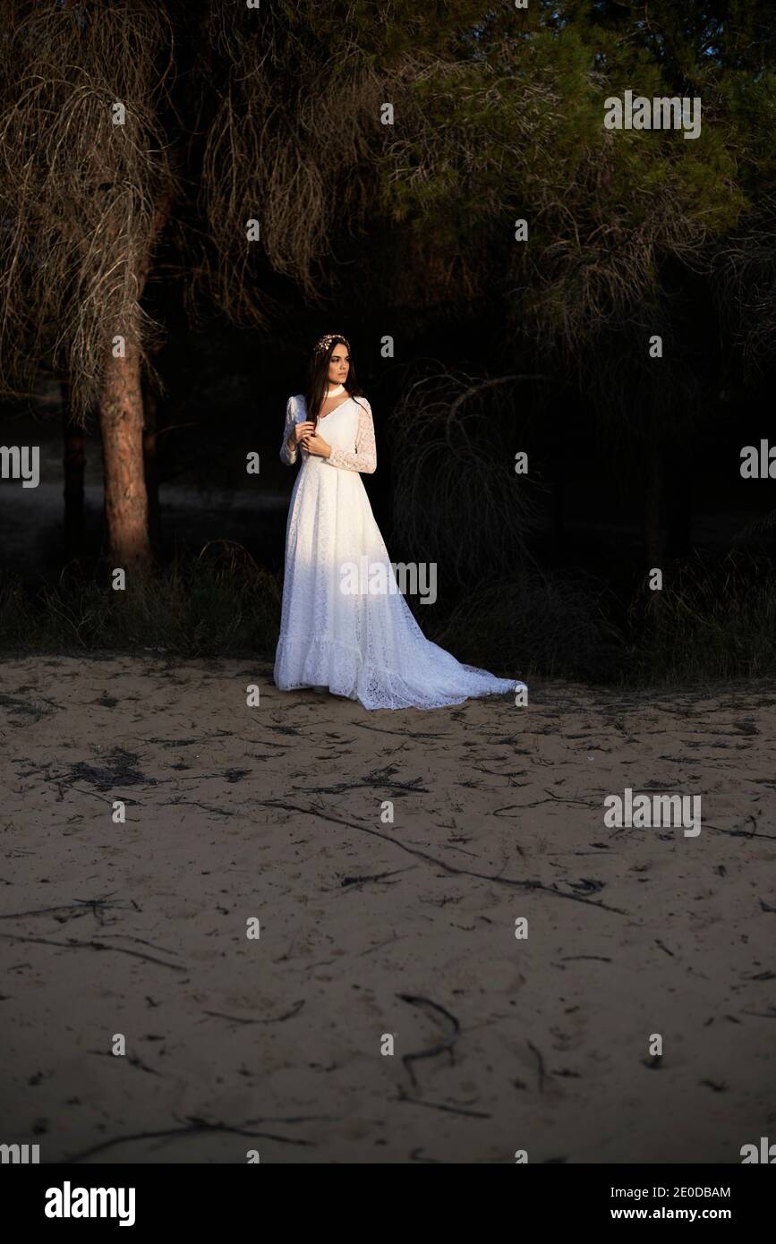 Vista laterale del corpo di felice elegante giovane femmina dentro boho abito da sposa maxi e corona in piedi su passerella di legno in boschi verdi e guardando lontano Foto Stock