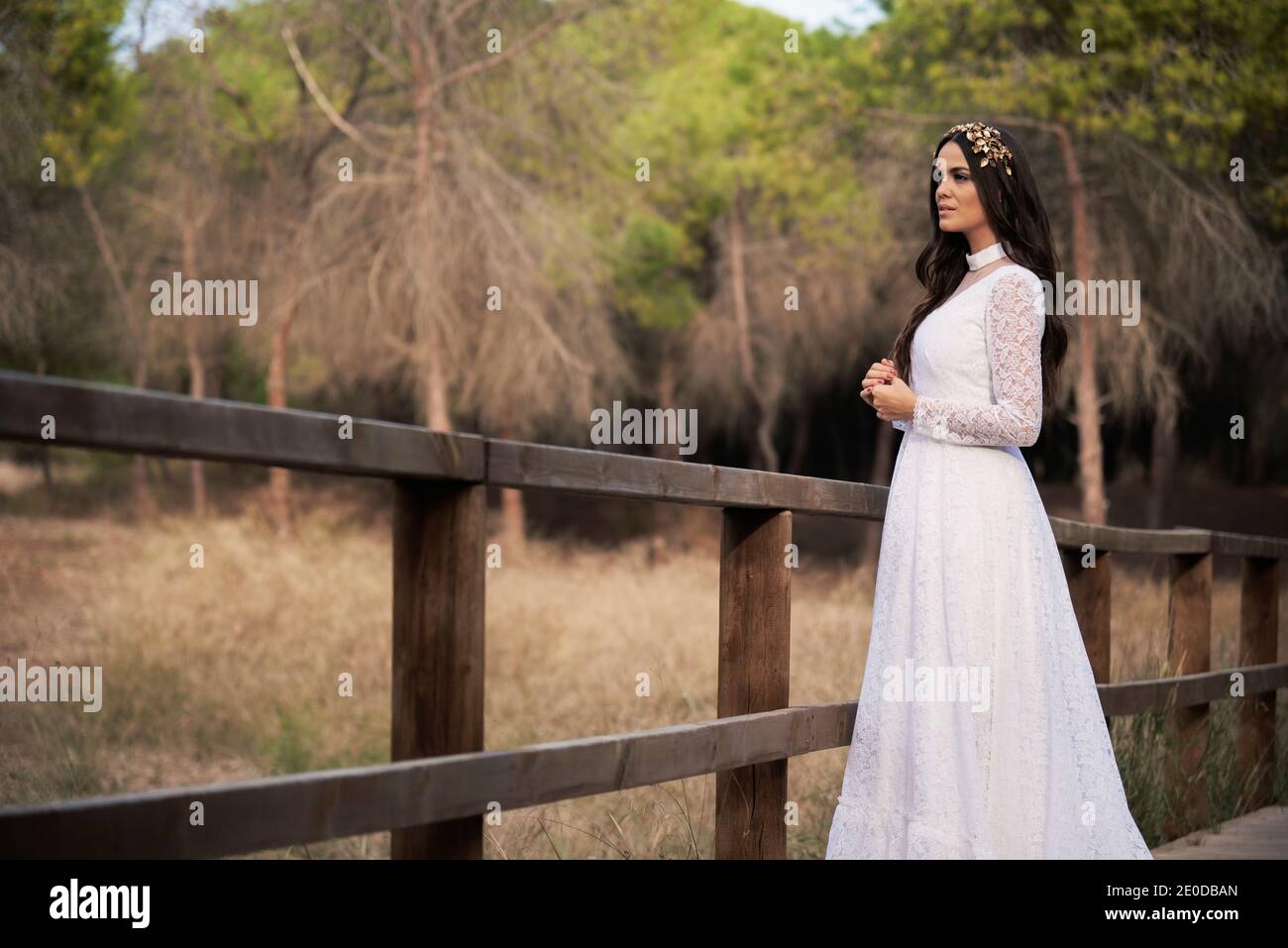 Vista laterale di felice elegante giovane femmina in boho nuziale maxi vestito e corona in piedi su passerella di legno in verde boschi e guardare lontano Foto Stock