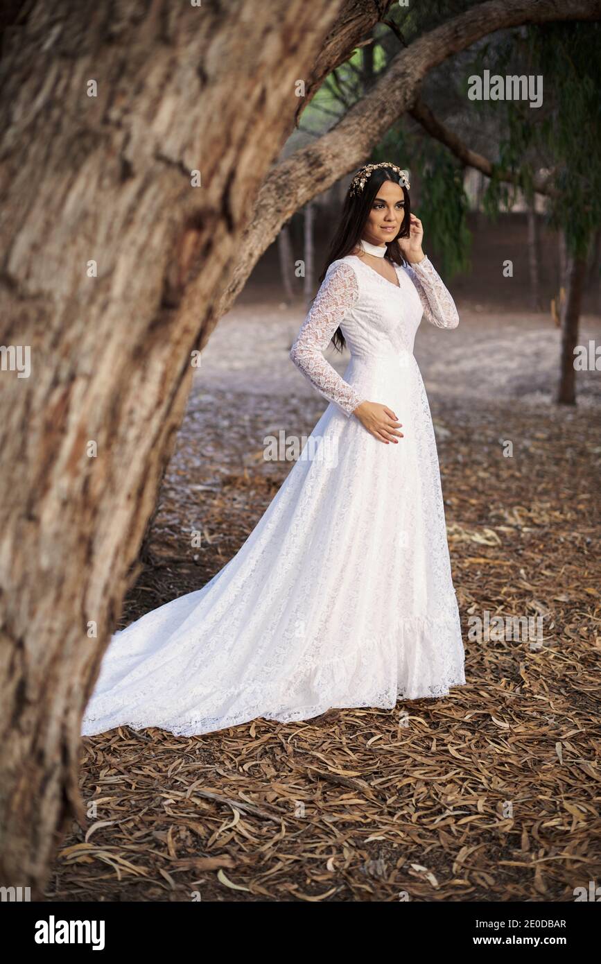 Vista laterale del corpo di felice elegante giovane femmina dentro boho abito da sposa maxi e corona in piedi su passerella di legno in boschi verdi e guardando lontano Foto Stock