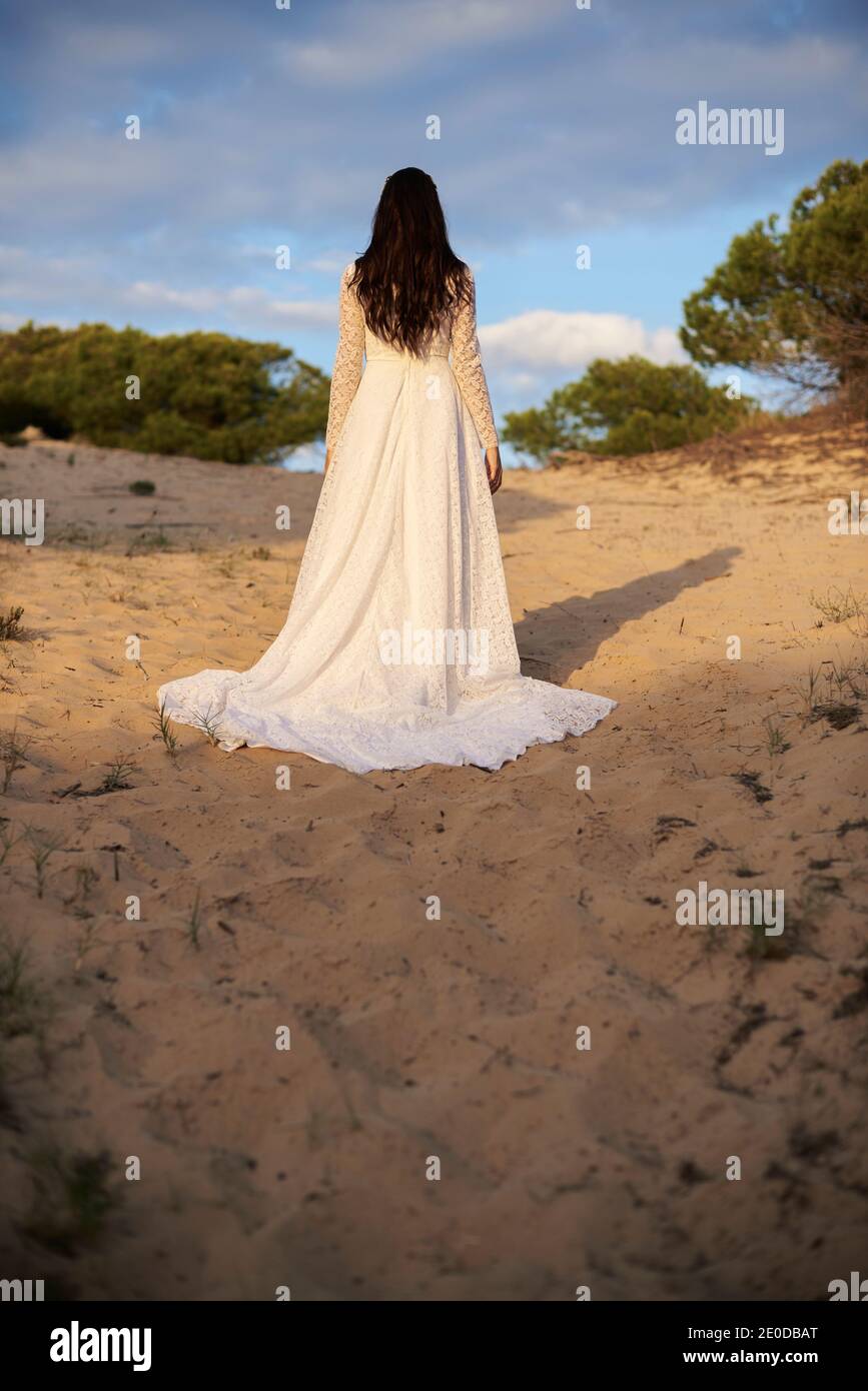 Vista posteriore di una bruna dai capelli lunghi irriconoscibile in maxi boho abito bianco in piedi su sabbia contro alberi verdi e. cielo nuvoloso Foto Stock