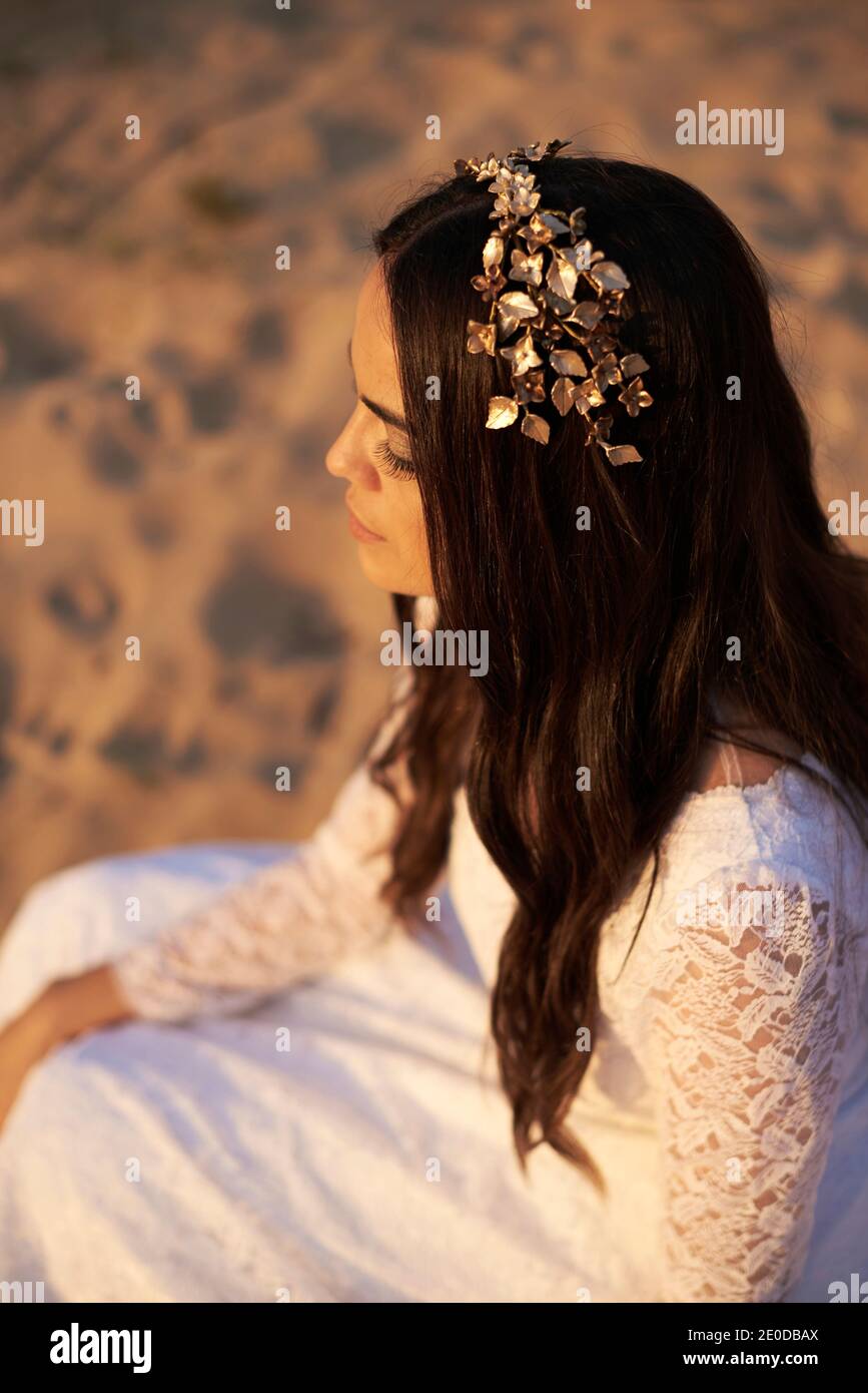 Da sopra vista laterale della giovane romantica brunetta bohémien in abito da sposa bianco e corona floreale dorata seduta sulla sabbia Foto Stock