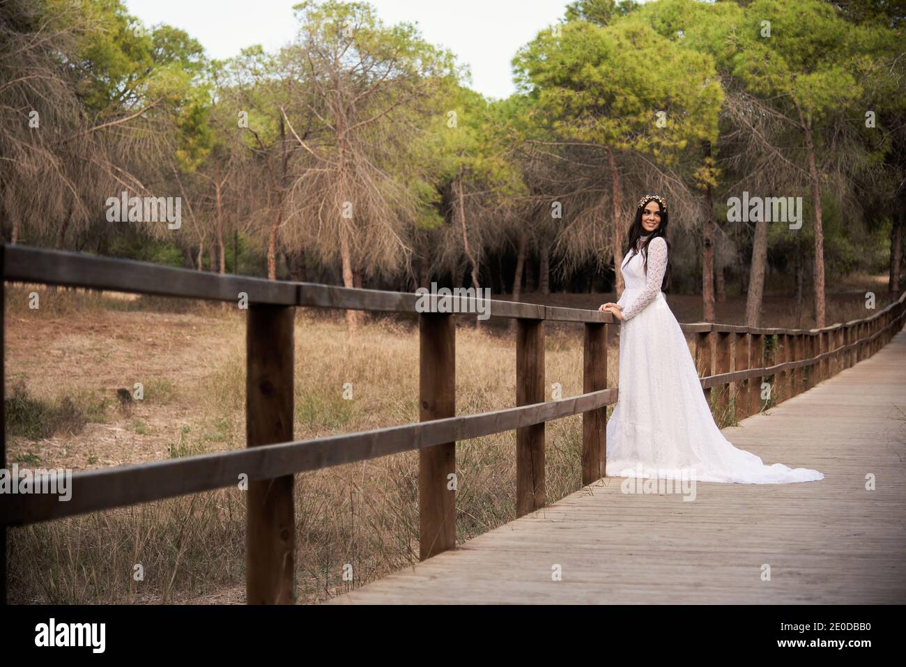 Vista laterale del corpo di felice elegante giovane femmina dentro boho abito da sposa maxi e corona in piedi su passerella di legno in legno verde e guardando la macchina fotografica Foto Stock