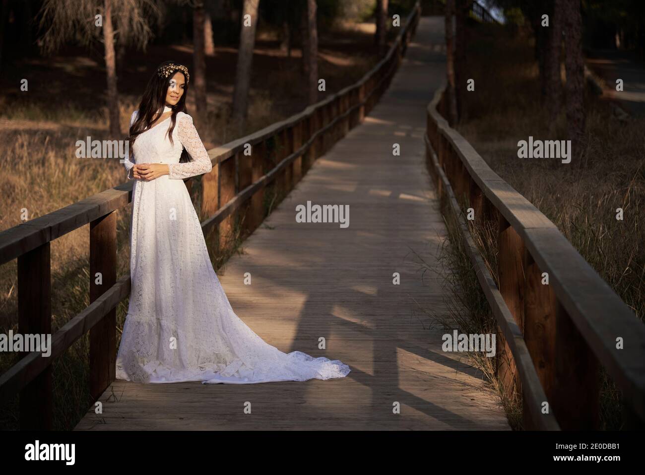Vista laterale del corpo di felice elegante giovane femmina dentro boho abito da sposa maxi e corona in piedi su passerella di legno in boschi verdi e guardando lontano Foto Stock