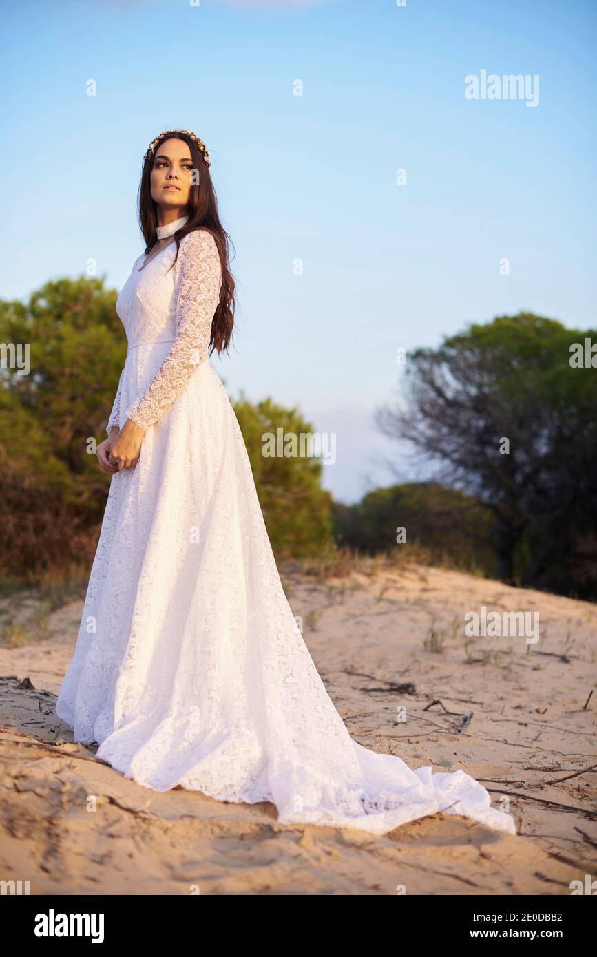 Vista laterale di elegante giovane femmina in boho bridal maxi vestitevi e cavatoie in boschi verdi e guardate via Foto Stock