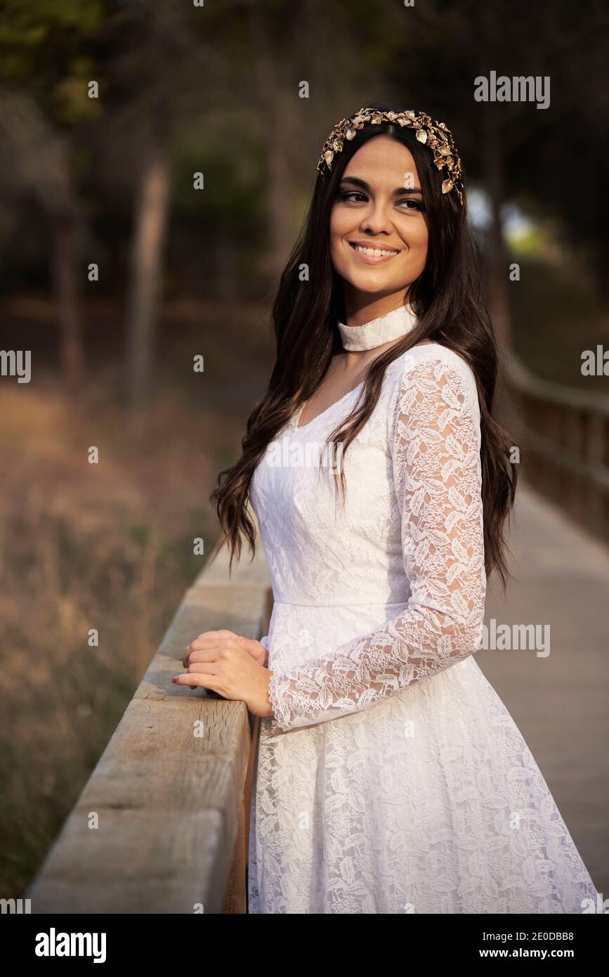 Vista laterale di felice elegante giovane femmina in boho nuziale maxi vestito e corona in piedi su passerella di legno in verde boschi e guardare lontano Foto Stock