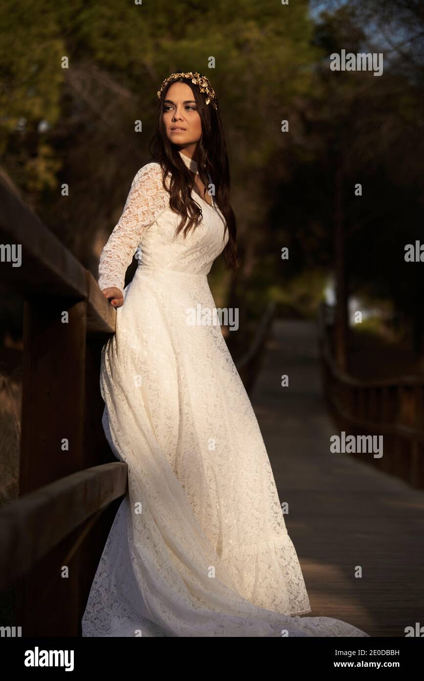 Vista laterale del corpo di felice elegante giovane femmina dentro boho abito da sposa maxi e corona in piedi su passerella di legno in boschi verdi e guardando lontano Foto Stock