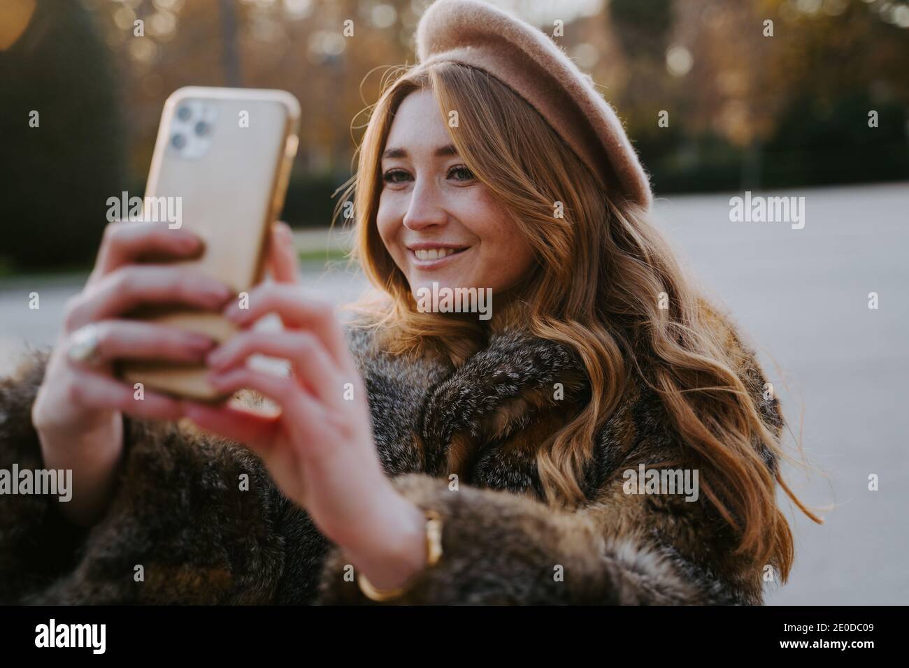 Affascinante donna in beret e cappotto caldo in piedi nel parco e portare selfie sullo smartphone mentre sorridi e ti godi il weekend Foto Stock