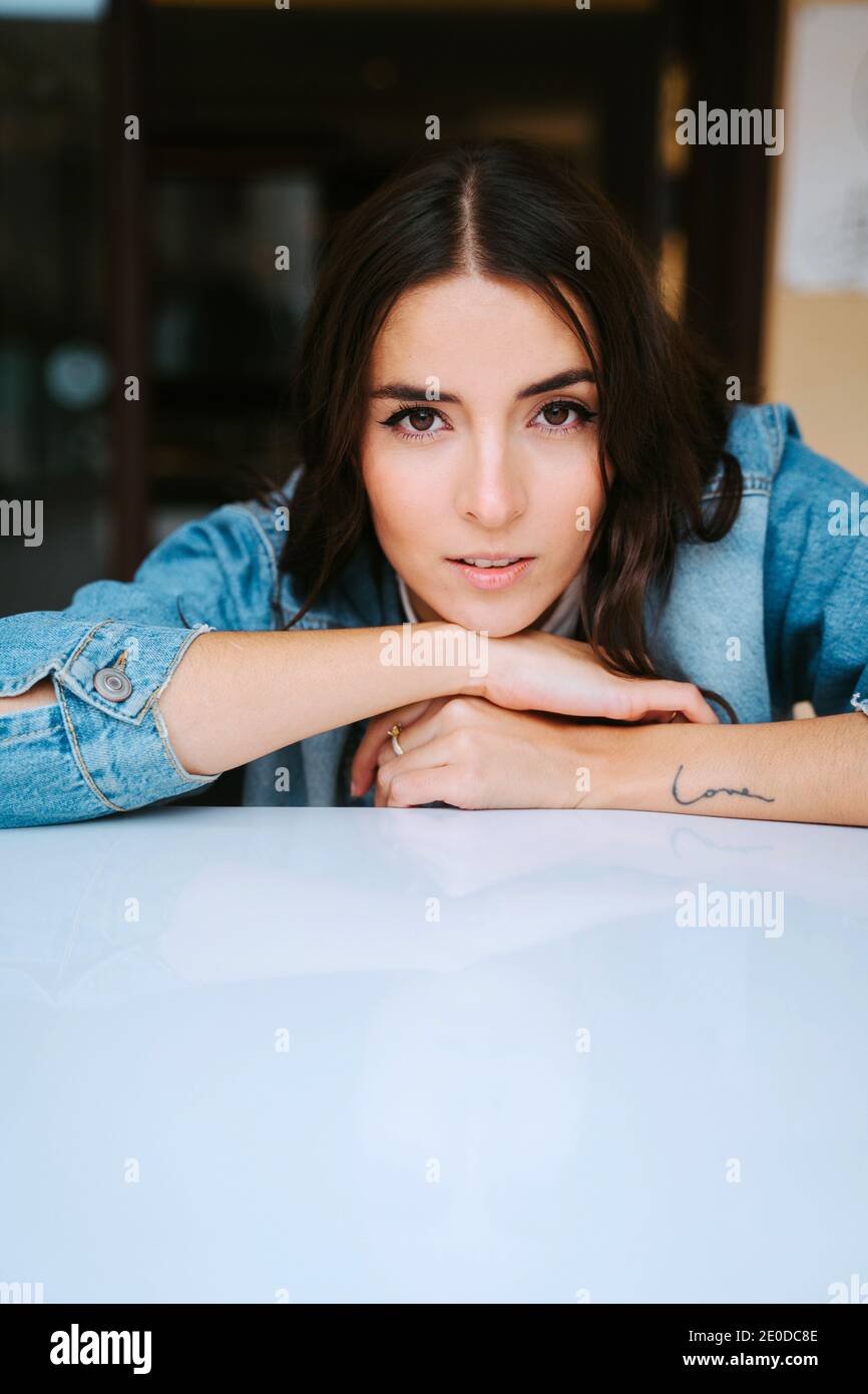 Bruna giovane calma positiva in denim e con tatuaggio sul braccio guardando la fotocamera mentre si riposa sul tavolo caffè Foto Stock