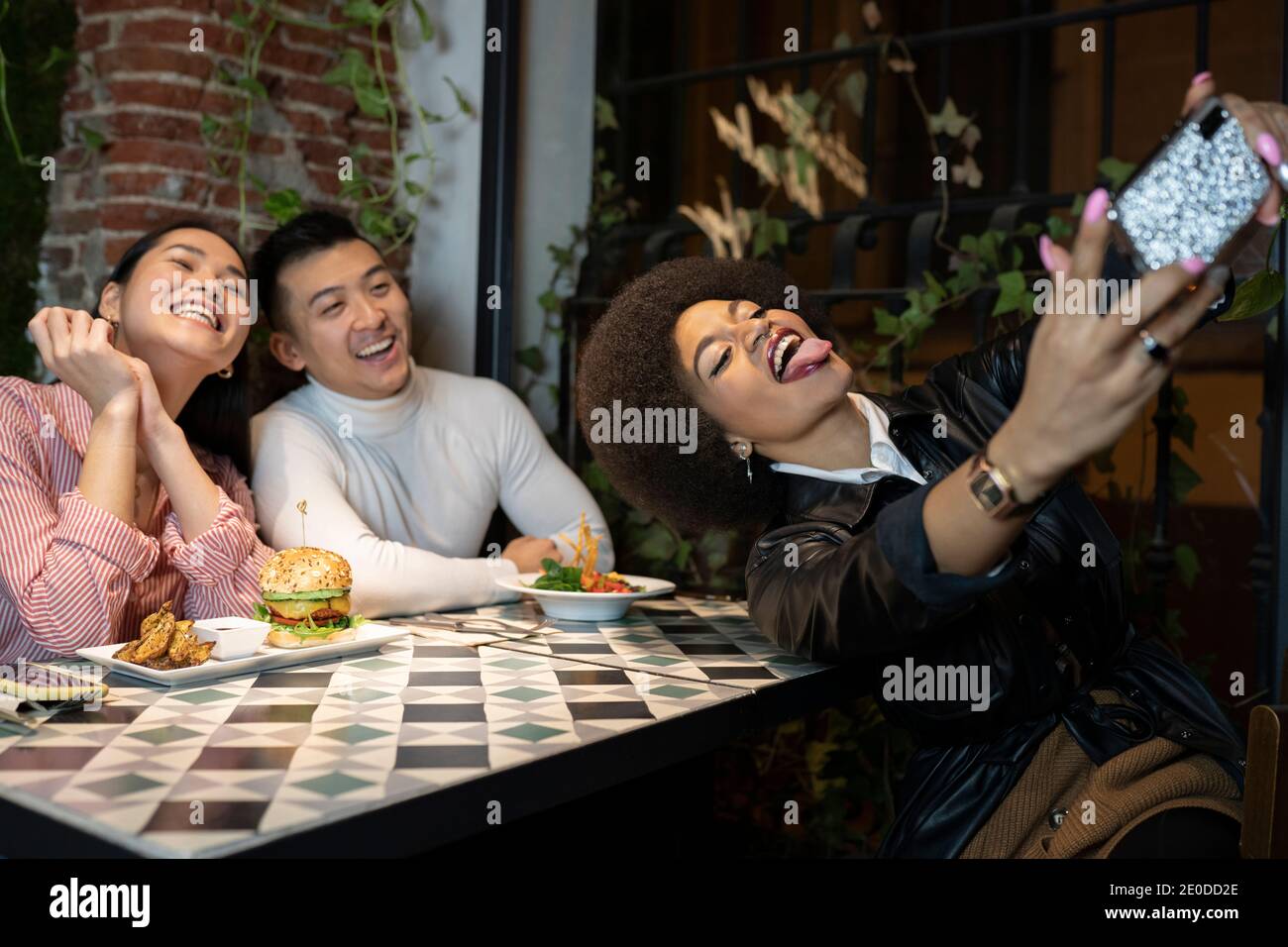 Allegra giovane afroamericana che mostra la lingua mentre prende selfie su smartphone con ridere fiends durante la cena in un accogliente ristorante Foto Stock