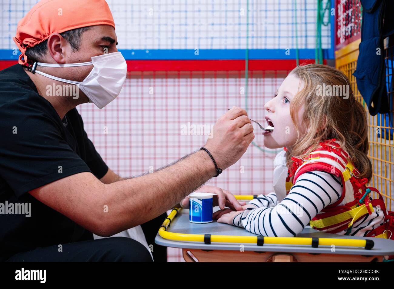 Vista laterale dello specialista maschile in maschera medica dando yogurt Per disabilitare la bambina WIT sindrome di Angelman mentre si ha la rottura durante lo sviluppo di attività Foto Stock
