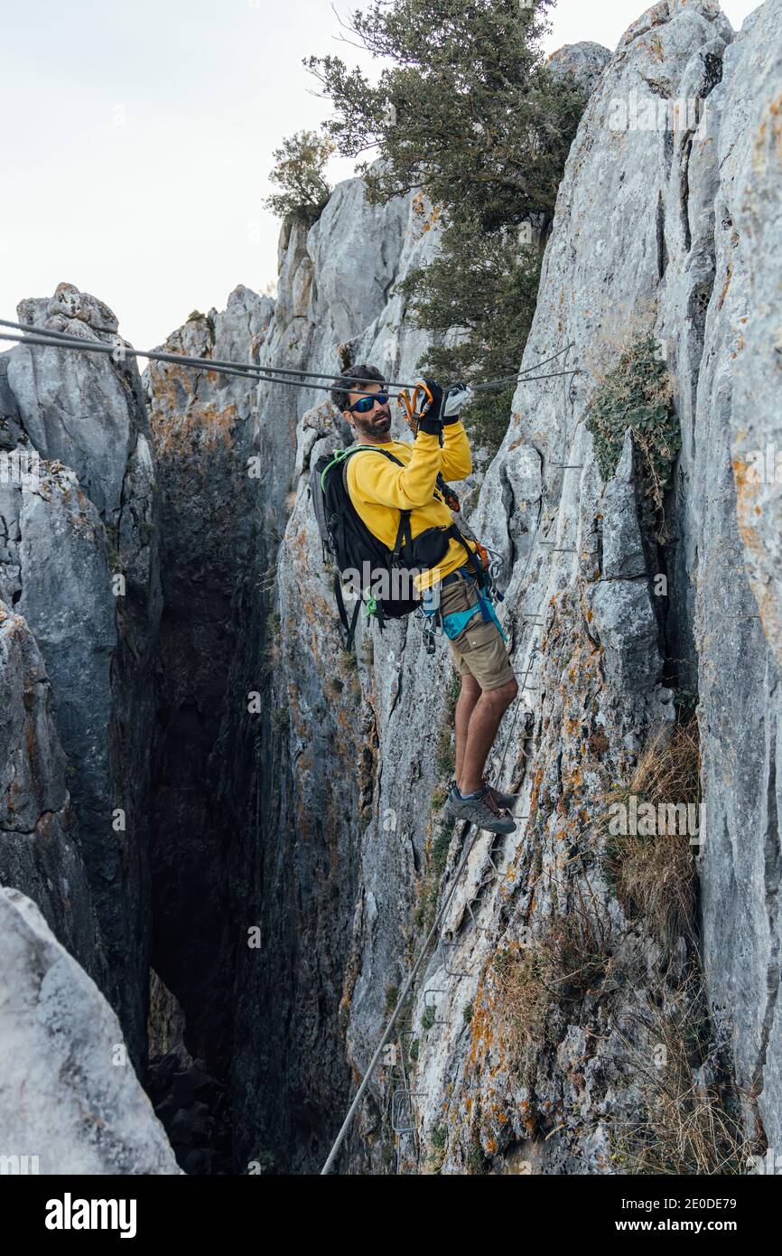 Vista laterale del corpo intero dell'alpinista maschile con zaino e. attrezzatura di sicurezza che si prepara ad attraversare burroni rocciosi su zipline mentre esercitandosi l'annuncio estremo Foto Stock