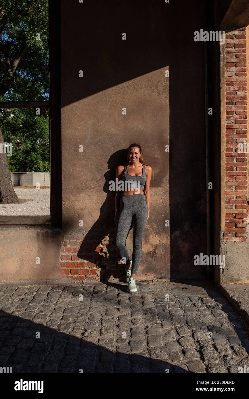 Allegro atleta in forma femminile in activewear appoggiata sulla parete di costruzione scortante e guardando la macchina fotografica mentre si è in piedi in città durante la formazione Foto Stock