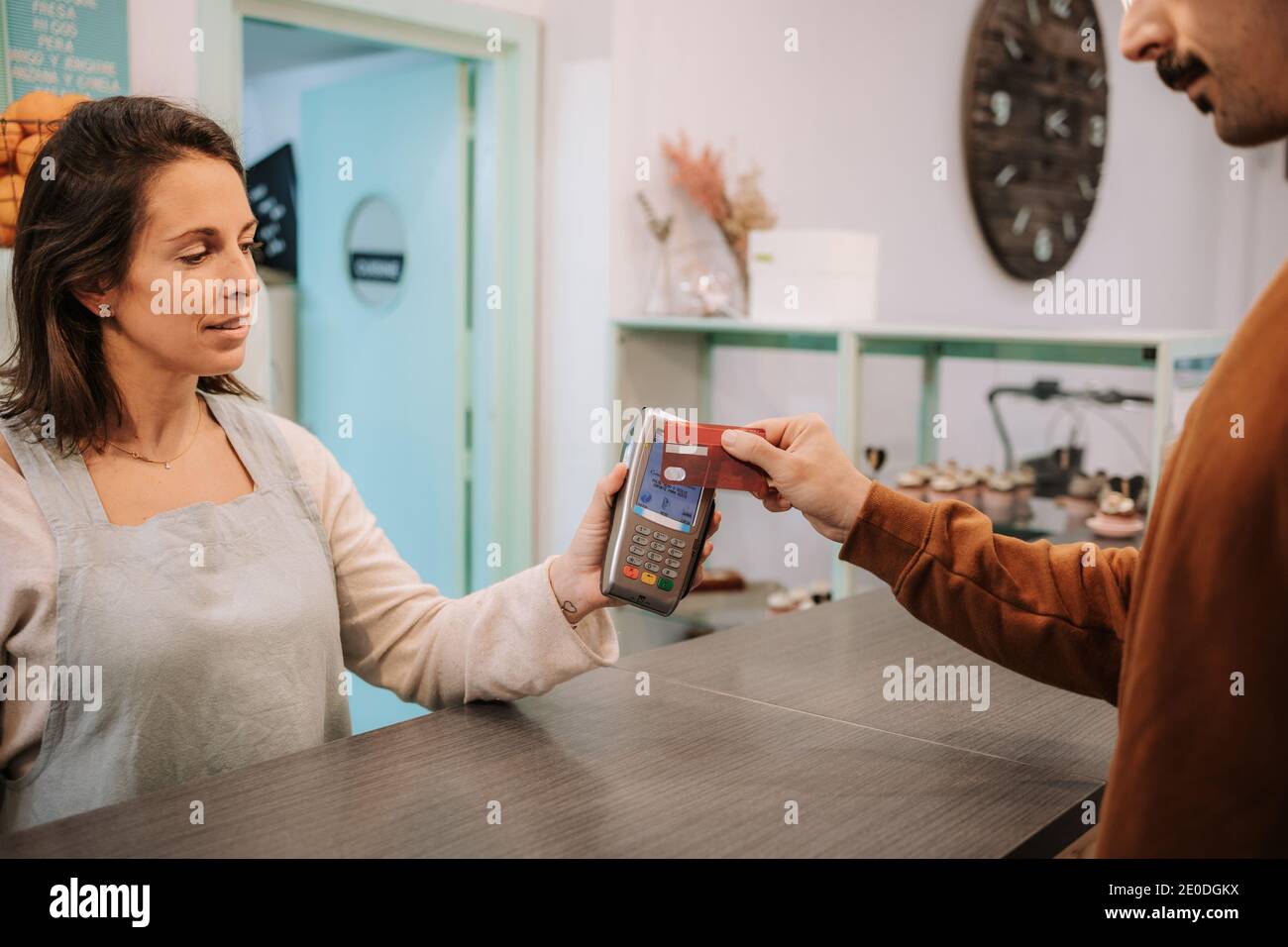 Venditore femminile positivo in piedi al banco e terminale di tenuta mentre Cliente che paga con carta di credito utilizzando la tecnologia NFC in pasticceria caffè Foto Stock