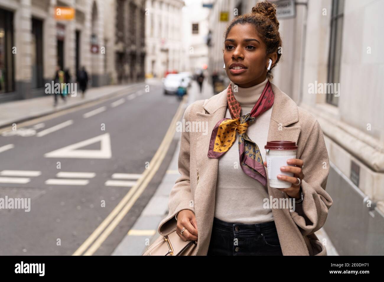 Giovane imprenditore afro-americano sicuro in outfit alla moda ascolto alla musica con gli auricolari wireless e bere il caffè da asporto whi Foto Stock