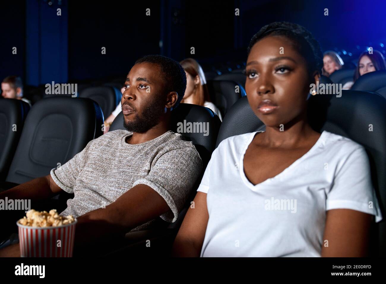 Vista laterale dell'uomo e della donna africani con volti forti che si godono il film. Fuoco selettivo di giovane coppia che guarda il film al cinema, seduto in comodi posti neri, indossando t-shirt casual. Foto Stock