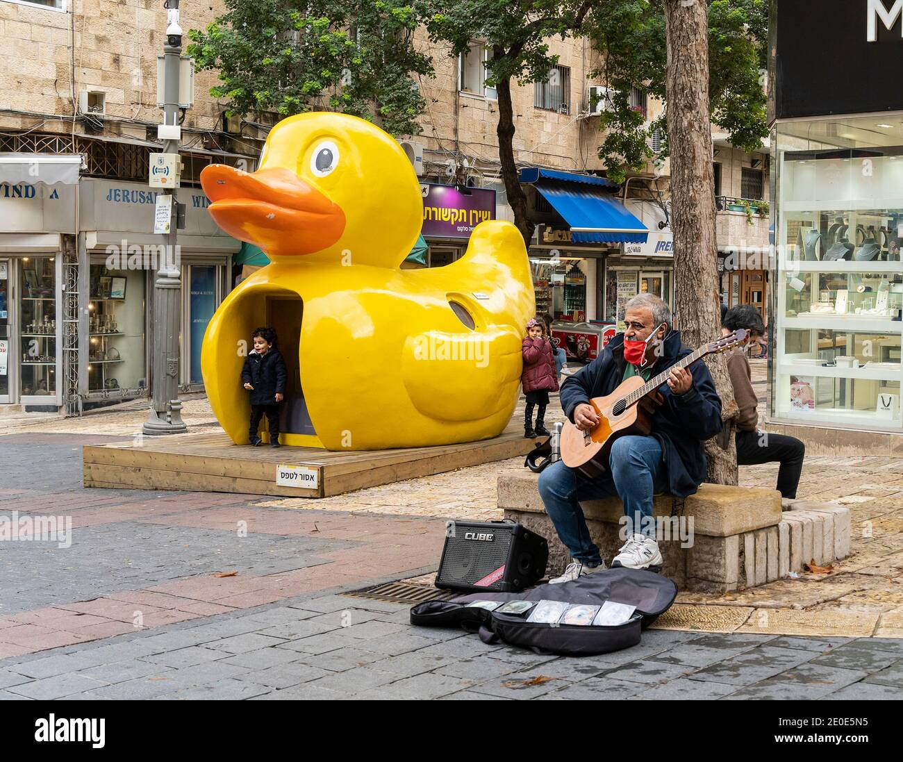 Gerusalemme, Israele - 17 dicembre 2020: Un uomo che suona la chitarra su una strada di Gerusalemme, mentre i bambini giocano intorno a una statua di un'enorme anatra di gomma. Foto Stock