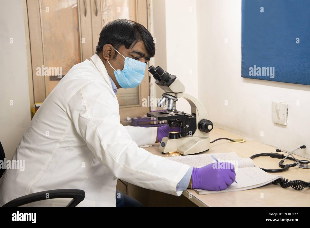 Medico o ricercatore asiatico indiano che indossa una maschera guardando attraverso un microscopio in un laboratorio. Uomo che prende nota o che fa un certo calcolo a. Foto Stock