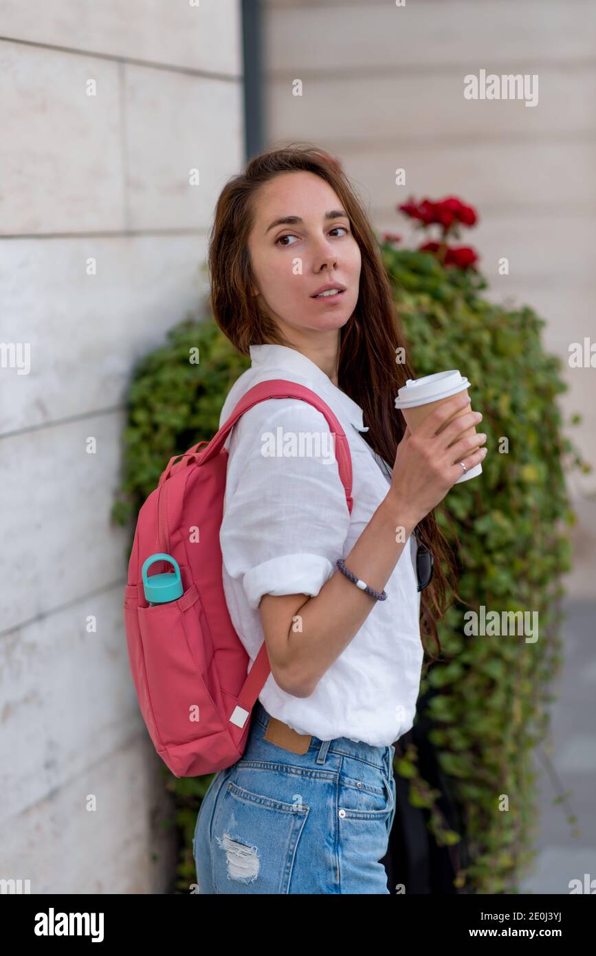 bella ragazza tiene una tazza di caffè e tè nelle sue mani, si gira in estate in città. Abbigliamento casual jeans camicia bianca e zaino rosa. Donna con Foto Stock