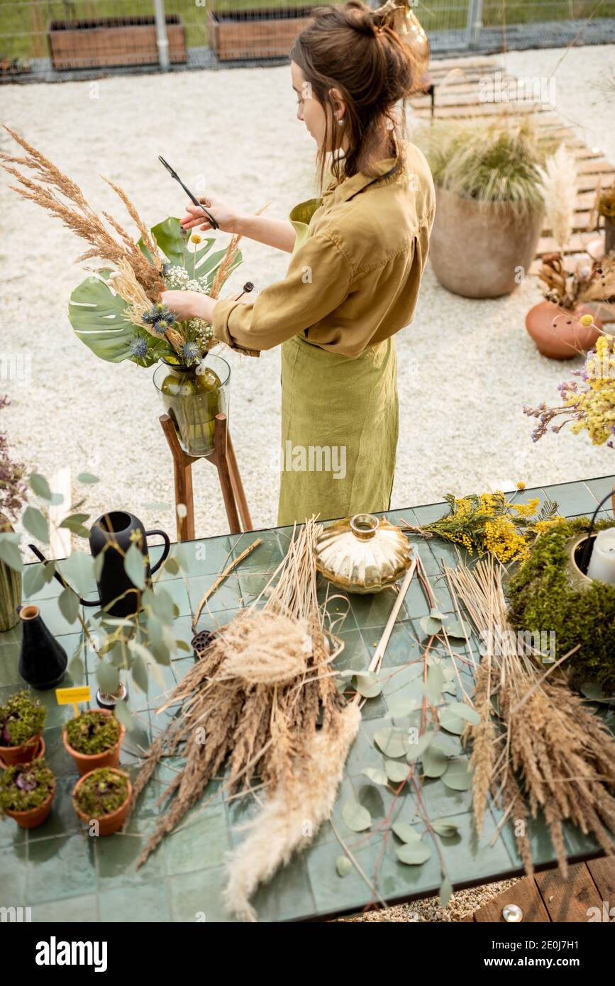 Giovane donna che fa composizioni di fiori secchi e freschi e di erbe in laboratorio all'aperto. Fiorista, giardiniere o decoratore che compone decorazione floreale Foto Stock
