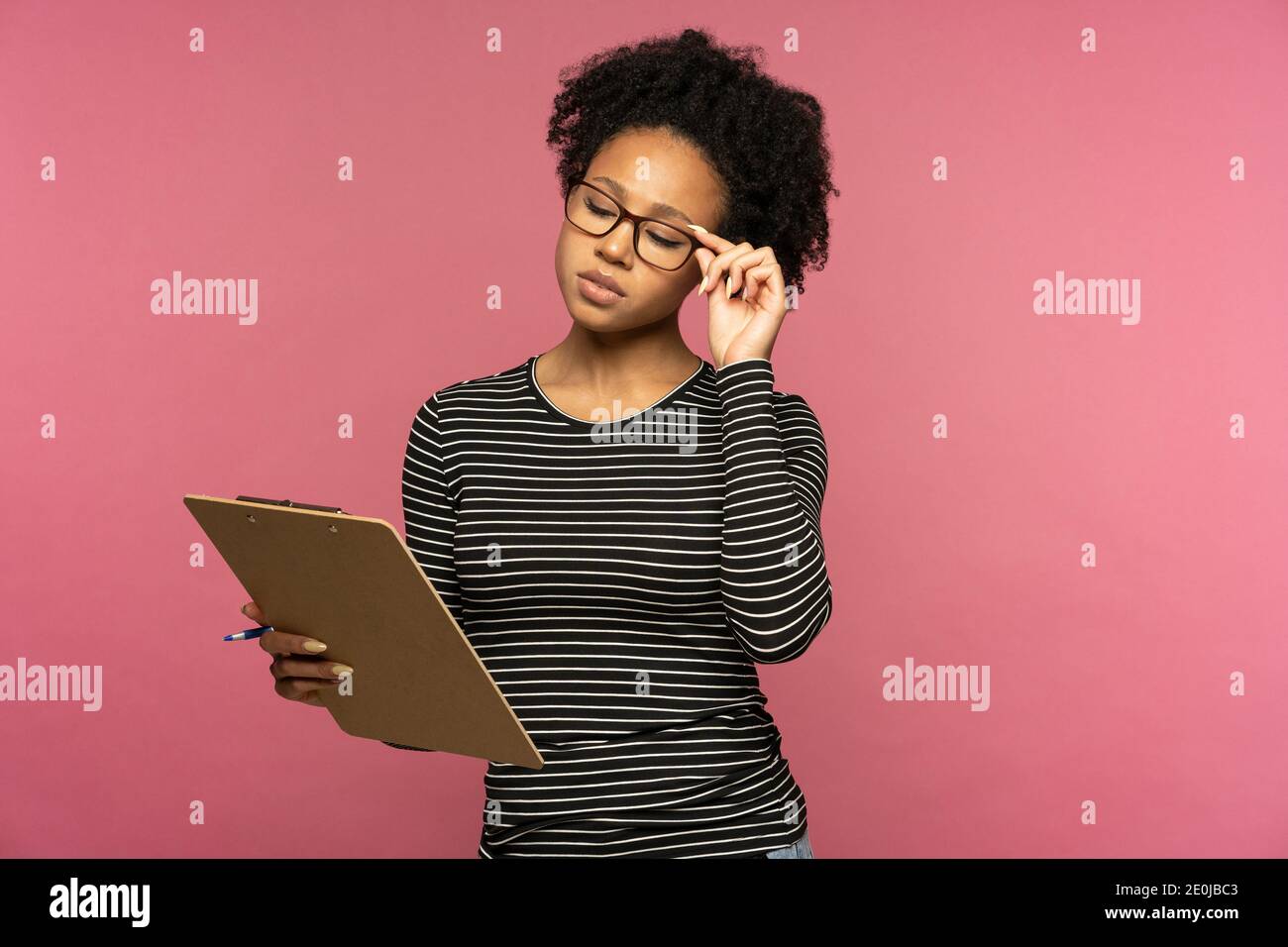 Donna studentesca afro-americana premurosa indossa occhiali tenendo cartella, fa note in documento, pensando, isolato sulla parete rosa dello studio. Concetto di pianificazione. Foto Stock