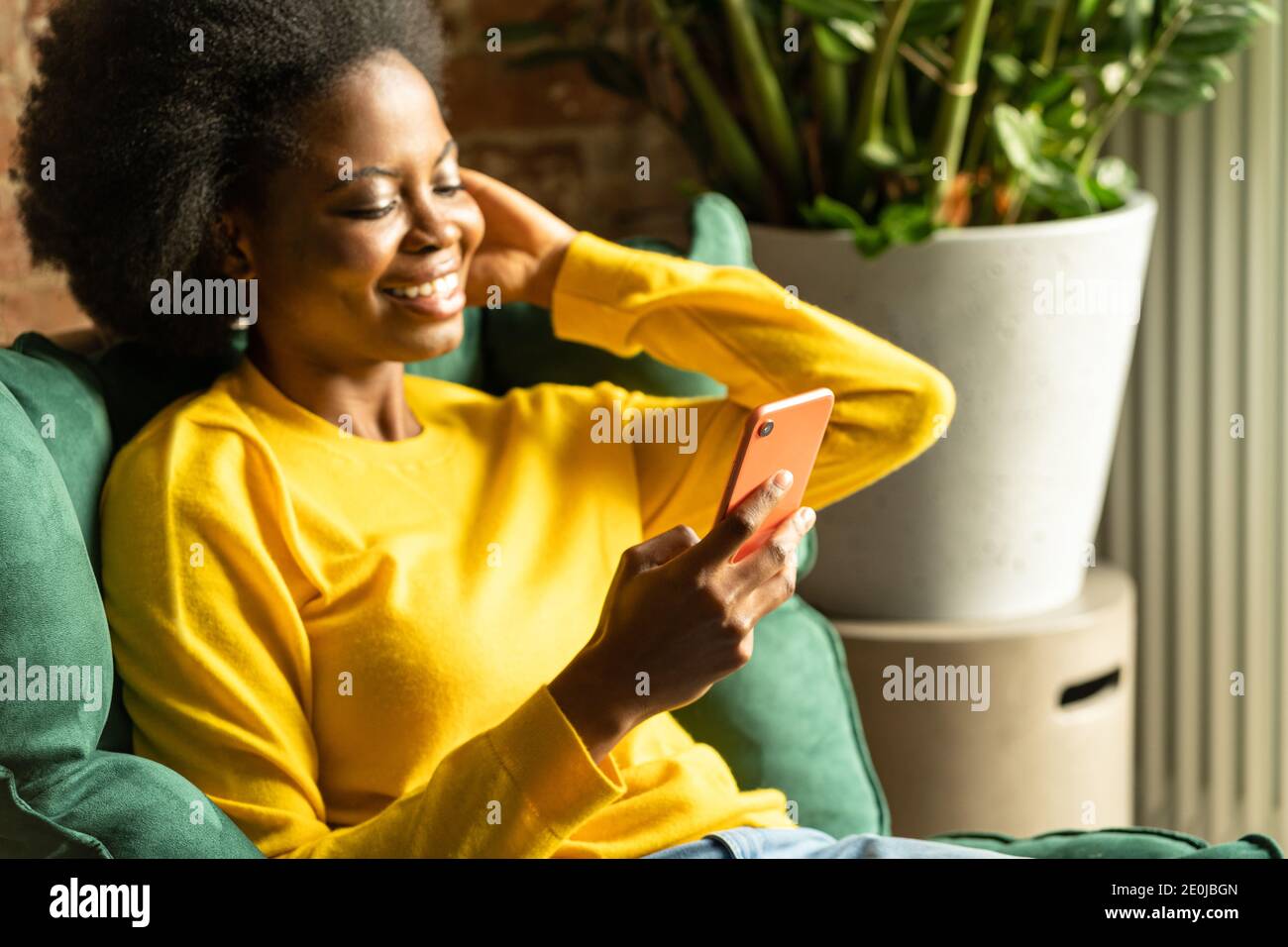 Primo piano della giovane donna millenaria afroamericana indossa un maglione giallo seduto su una sedia verde, riposando, utilizzando smartphone, digitando messaggio Foto Stock