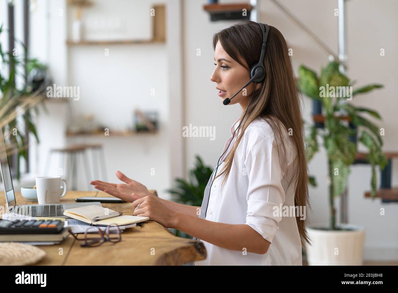 Vista laterale della donna caucasica freelancer focalizzata in auricolari seduti a tavola, parlando in video chat, lavorando a distanza dall'ufficio domestico. Lavoro a distanza. Foto Stock