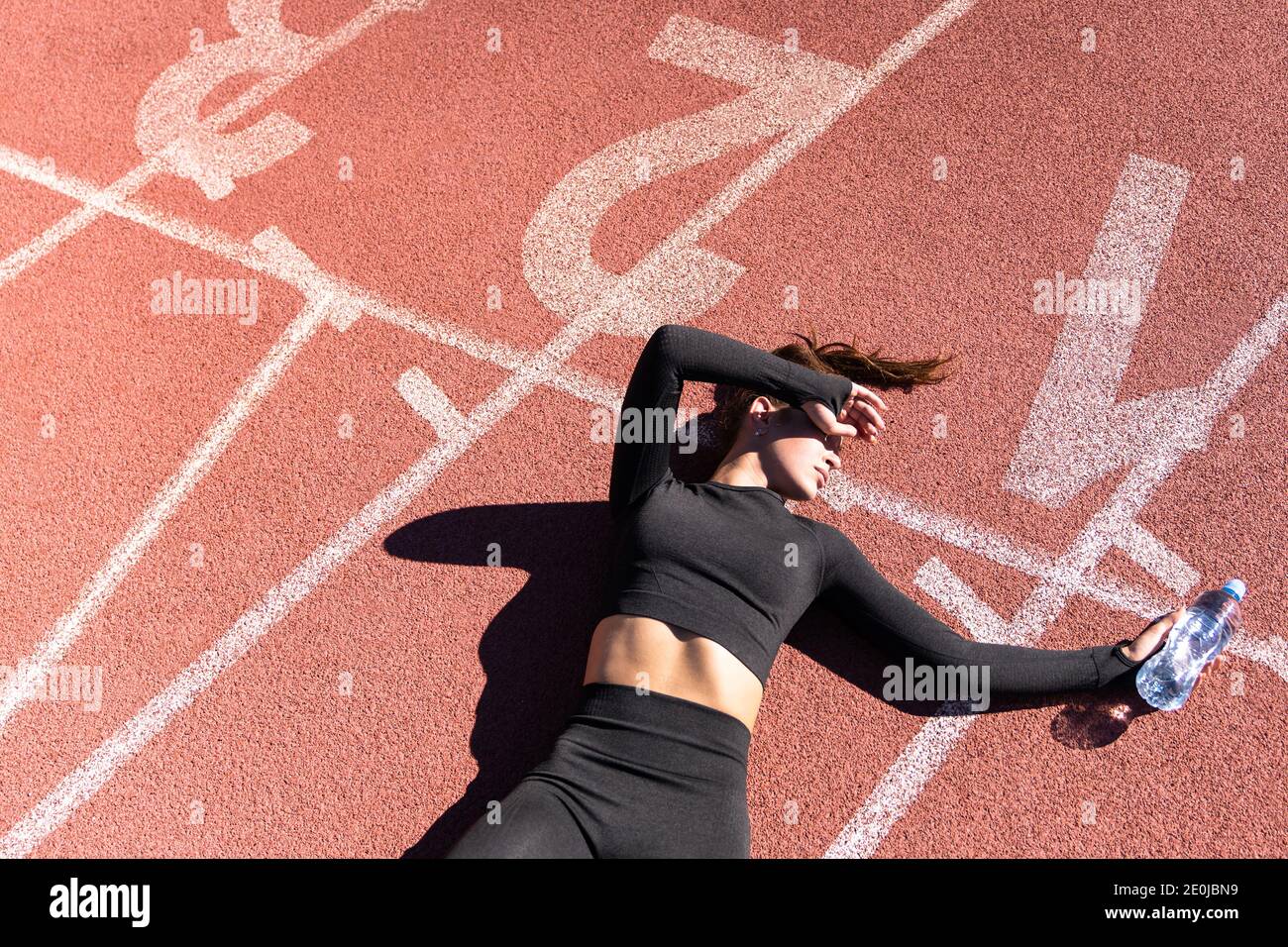 Vista dall'alto di una donna stanca in forma sportiva che riposa dopo l'allenamento o che corre su uno stadio di gomma tapis roulant, tenendo una bottiglia d'acqua, facendo una pausa du Foto Stock