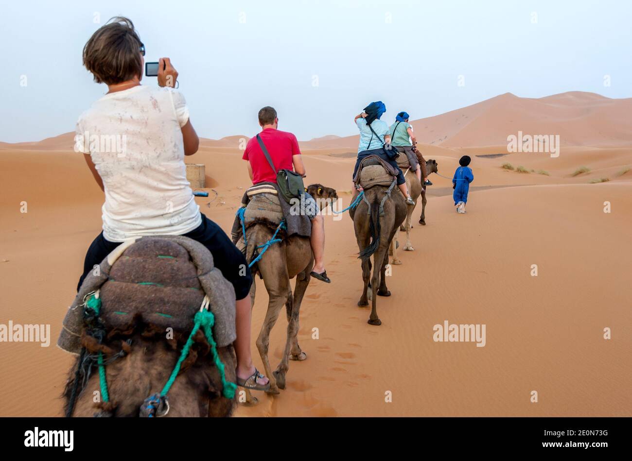Turisti stranieri cavalcano cammelli nel deserto al tramonto a Merzouga, che si trova sul bordo di Erg Chebbi (il Mare di sabbia) in Marocco. Foto Stock