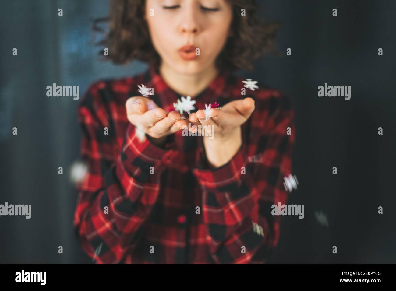 Attraente giovane donna in abito rosso plaid che soffia sulle sue mani con fiocchi di neve, umore invernale Foto Stock