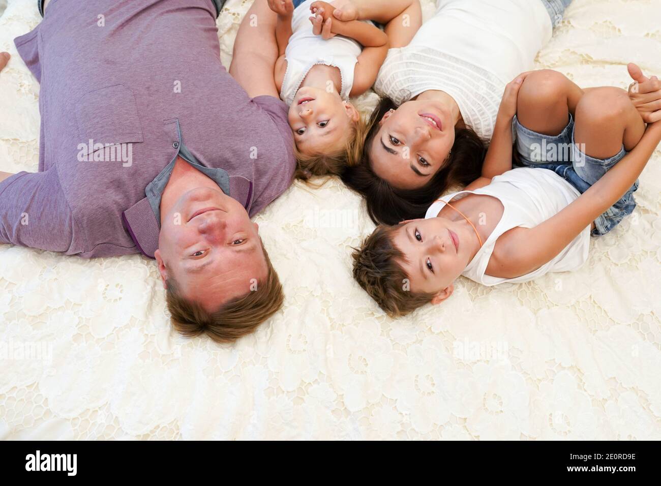 Vista dall'alto della bella giovane madre, del padre e dei loro figli guardando la macchina fotografica e sorridendo mentre si stende sul letto testa a testa Foto Stock