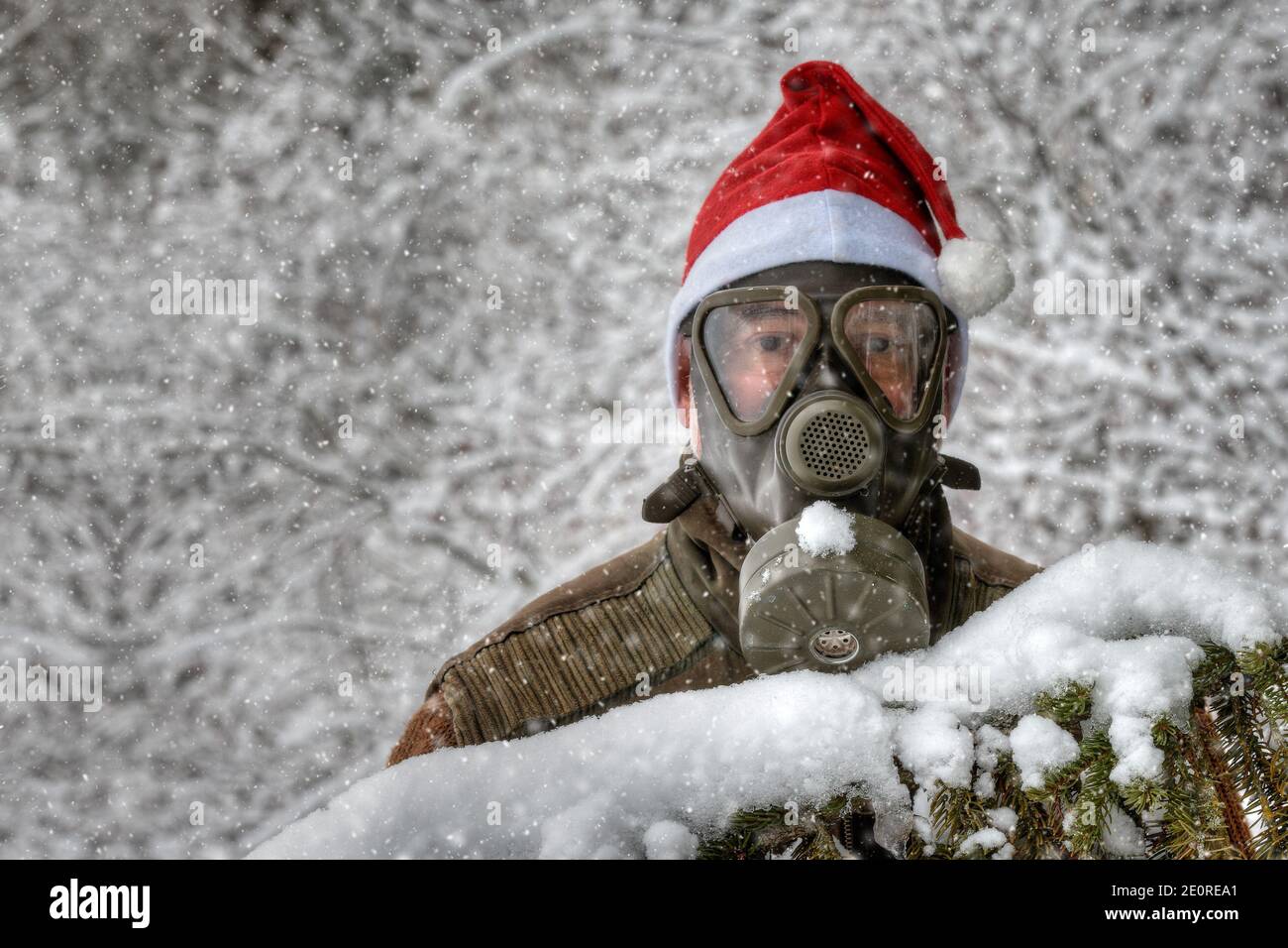 Dietro un ramo innevato di abete si trova un uomo con una maschera a gas e un cappello rosso Santa in una nevicata nel paesaggio invernale. Foto Stock