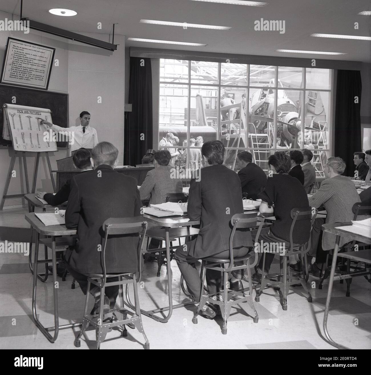 Anni '50, tirocinanti storici, giovani ingegneri o apprendisti in classe, Aviazione. Un segno sopra la lavagna dice, 'il valore della scienza dell'artigianato', che dichiara, 1; sviluppare la capacità di analizzare un problema o una procedura di officina......e continua ad elencare i numerosi altri benefici. In distanza, il pavimento della fabbrica e un piccolo aeroplano propellered. Foto Stock