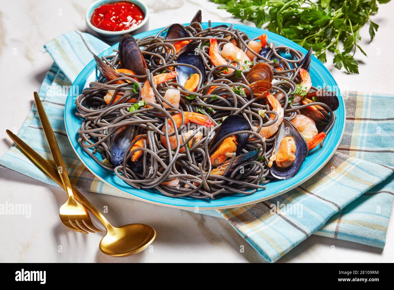 Pasta con inchiostro nero calamaro e frutti di mare: Cozze e gamberetti con vino bianco e salsa all'aglio su un piatto blu con posate dorate su uno sfondo di marmo bianco Foto Stock