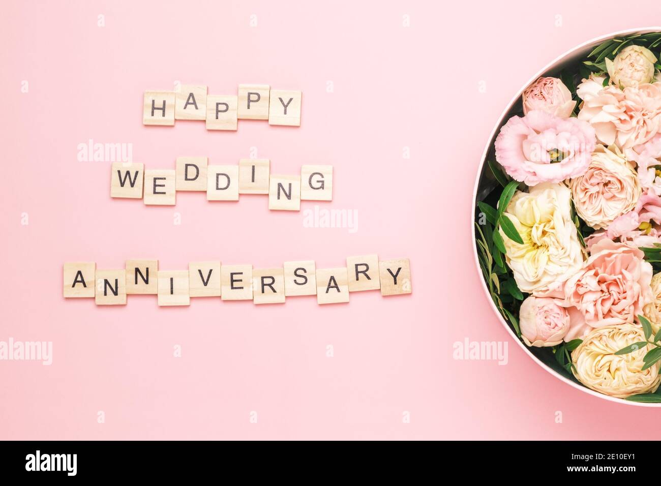 Felice anniversario di matrimonio scritta sulle piazze di legno con lettere sullo sfondo rosa e grande scatola rotonda con fiori rossi e rose. Foto Stock