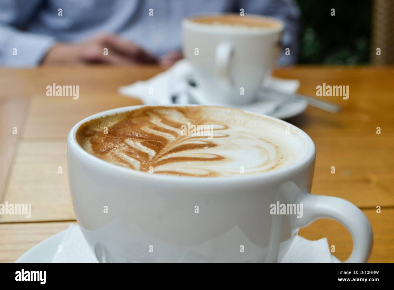 Due tazze di cappuccino o latte di caffè aromatico su un tavolo di legno. Sullo sfondo, una persona parzialmente visibile è seduta al tavolo. Foto Stock