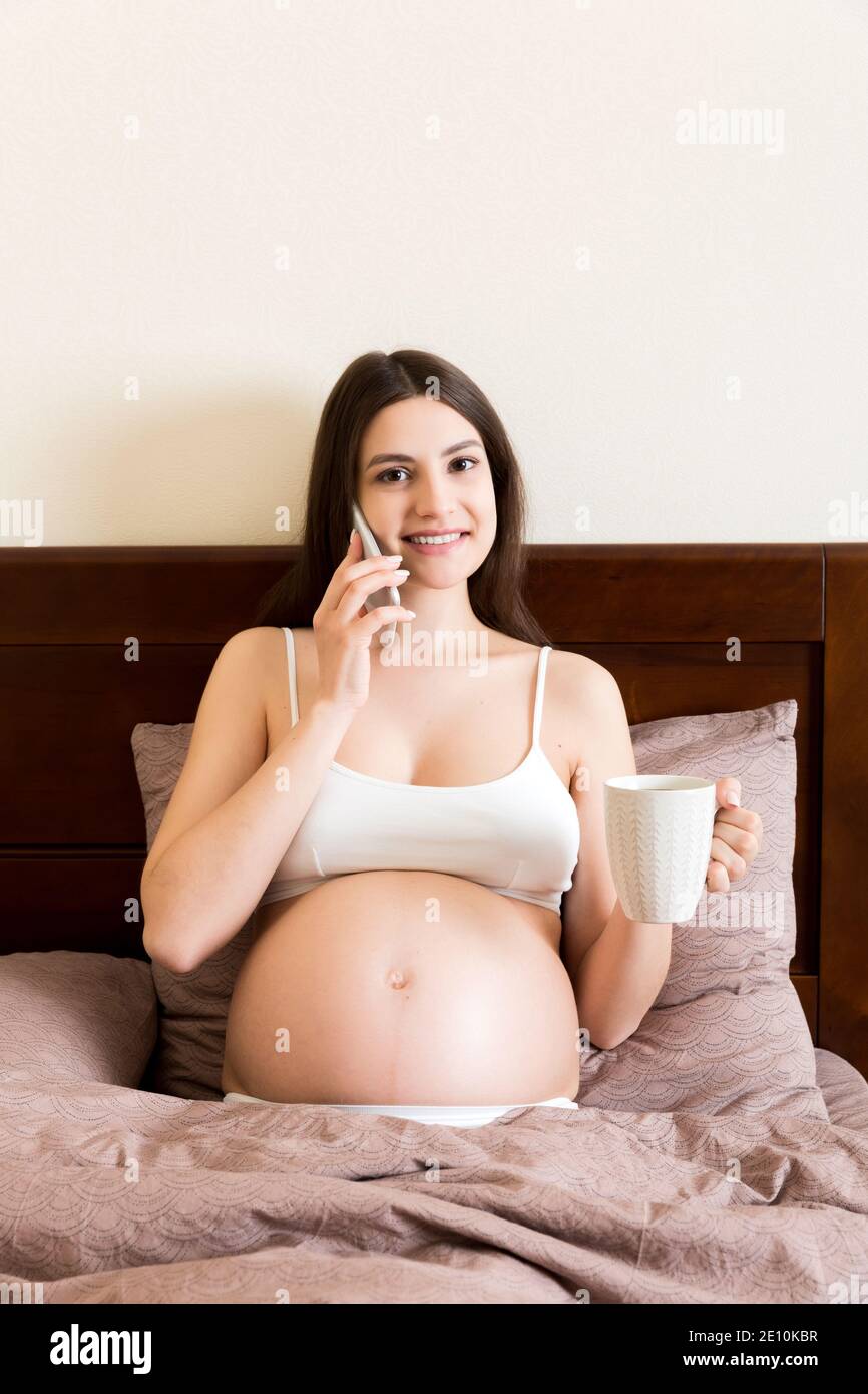 giovane donna incinta seduto a letto e bere caffè e parlare messaggio a casa. concetto di maternità. Foto Stock