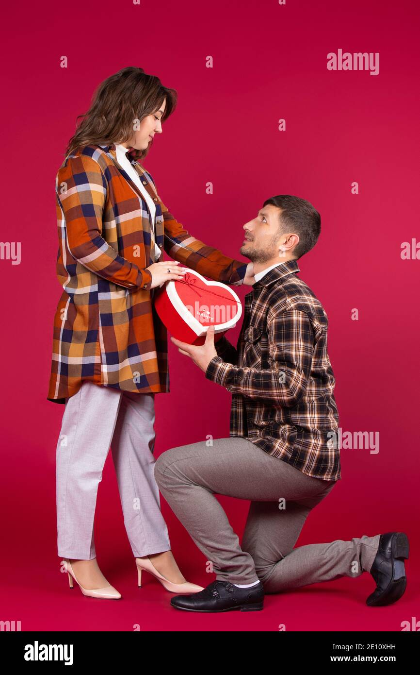 San Valentino. Un uomo in ginocchio dà una scatola regalo rossa a forma di cuore. Sfondo rosso. Foto di alta qualità Foto Stock