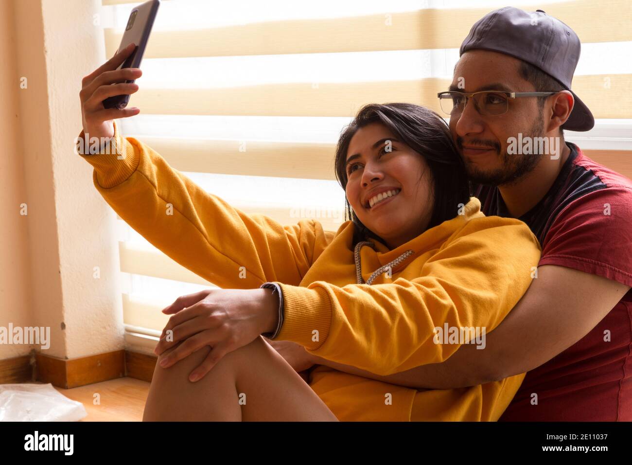 coppia che gioca e prende un selfie, due persone con abiti industriali per dipingere, sposa e sposo sorridendo seduto di fronte. una finestra Foto Stock