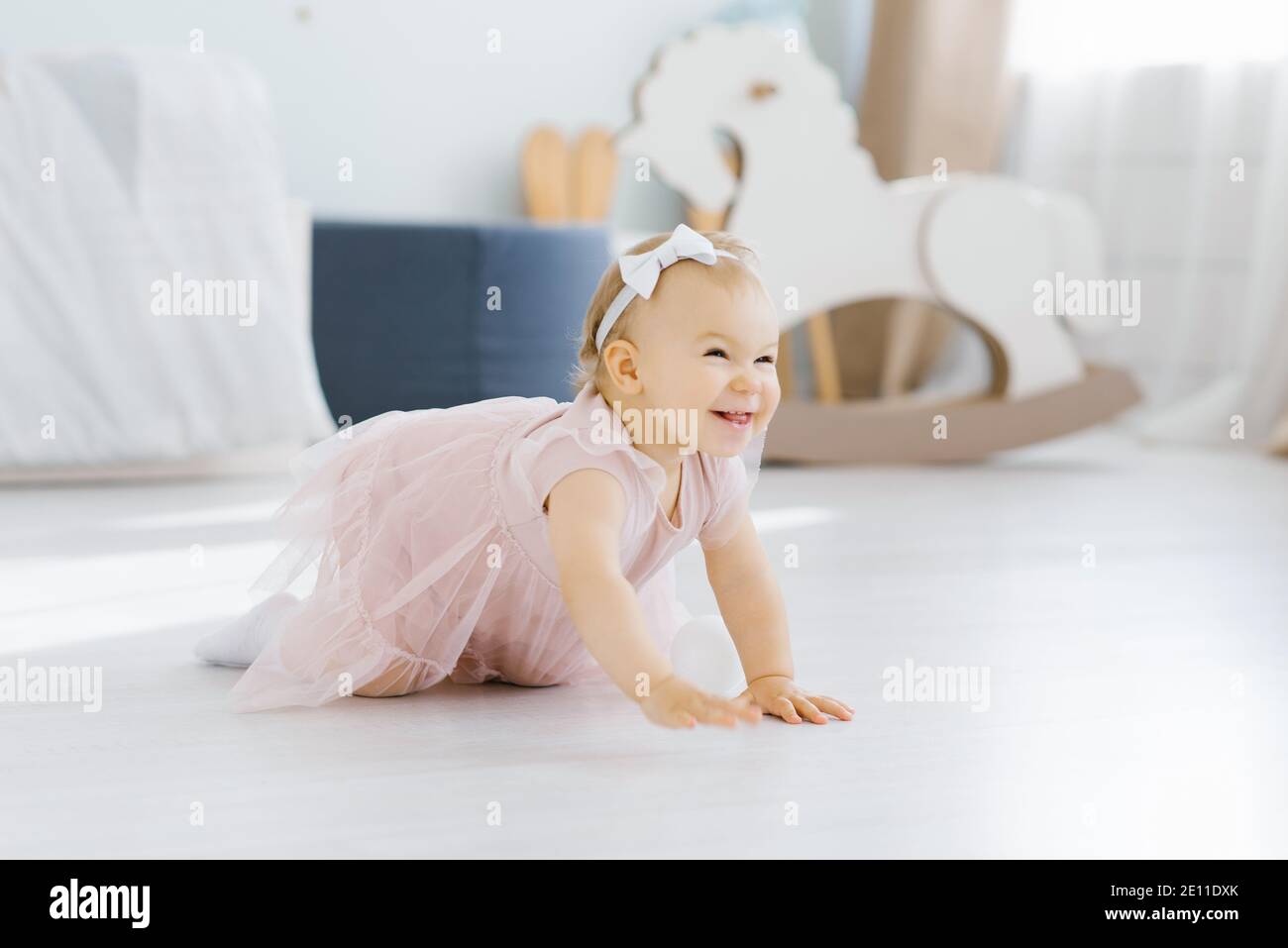 Una bella bionda con gli occhi blu di un anno in un l'abito rosa striscia su tutte le farine nella stanza dei bambini sopra il pavimento e sorride fervente Foto Stock