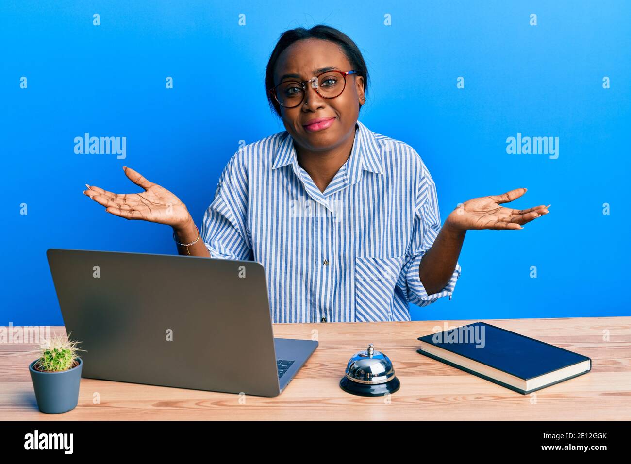 Giovane donna africana che lavora alla reception dell'hotel usando il computer portatile senza indizi e confuso l'espressione con le braccia e le mani sollevate. Concetto di dubbio. Foto Stock