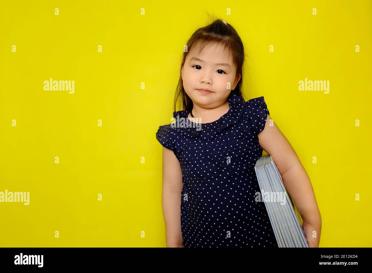 Una giovane ragazza asiatica carina è portare un grande libro di testo sotto il braccio, sorridendo, preparandosi per l'ora della storia a scuola. Sfondo giallo brillante. Foto Stock